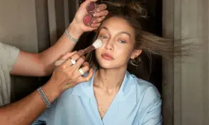 En makeupartist applicerar foundation med en borste på en kvinna med blont hår. Två små burkar med rouge syns i handen.