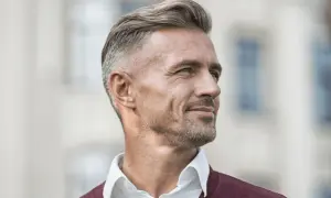 En man med grått hår, klädd i en vit skjorta och en vinröd tröja, tittar bort med ett leende.