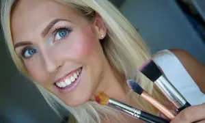 Blond kvinna med blå ögon ler och håller sminkborstar. Hon har precis applicerat rouge för en frisk lyster.
