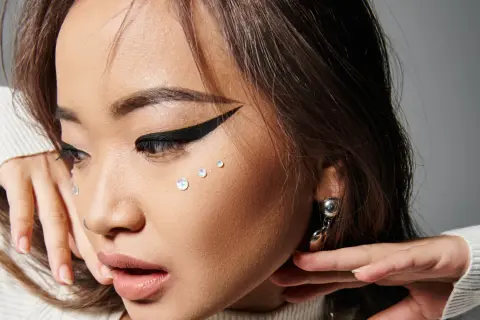 En närbild på en ung kvinna med en dramatisk eyeliner vinge, glittrande kristaller på kinden och en silverfärgad örhänge.