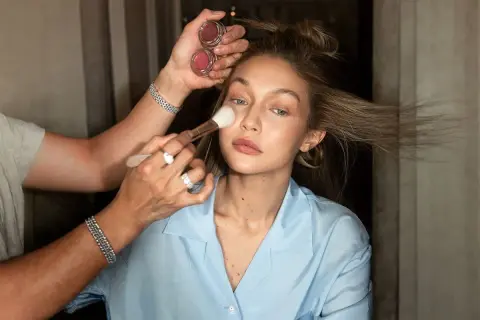 En makeupartist applicerar foundation med en borste på en kvinna med blont hår. Två små burkar med rouge syns i handen.