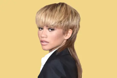 Zendaya visar upp en modern mullet med blont hår och en svart kavaj mot en gul bakgrund.