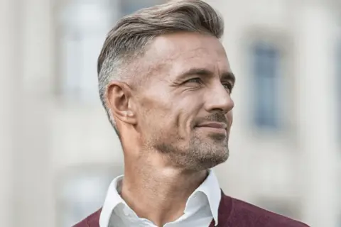 En man med grått hår, klädd i en vit skjorta och en vinröd tröja, tittar bort med ett leende.