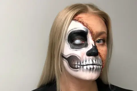 En kvinna med en otrolig ansiktsmålning för halloween. Halva ansiktet är målat som ett skelett med en vit pupill, medan andra halvan visar en realistisk, trasig hudeffekt.