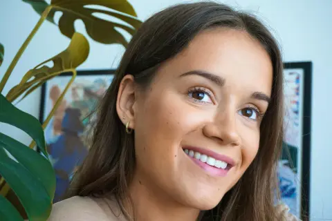 En kvinna ler med en naturlig no makeup makeup-look. En grön Monstera-växt skymtar i förgrunden.