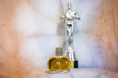 En gyllene flaska med naturliga parfymer, "LenaBlum Out of Woods", står framför en Oscarstatyett.