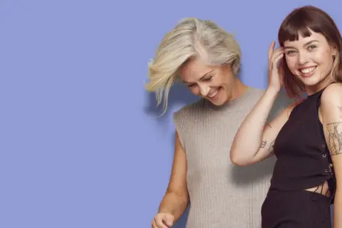 Två kvinnor ler mot kameran. Den ena har en kort rufsig frisyr dam och grått hår, den andra har mörkt hår och tatueringar.
