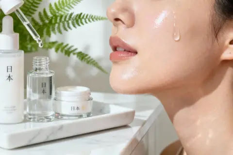 En droppe serum faller mot en glänsande hud, omgiven av japanska skönhetsprodukter. J beauty för en strålande lyster.