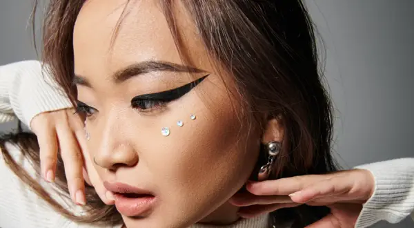 En närbild på en ung kvinna med en dramatisk eyeliner vinge, glittrande kristaller på kinden och en silverfärgad örhänge.