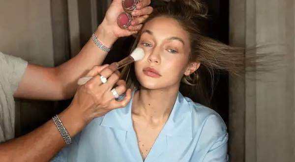 En makeupartist applicerar foundation med en borste på en kvinna med blont hår. Två små burkar med rouge syns i handen.
