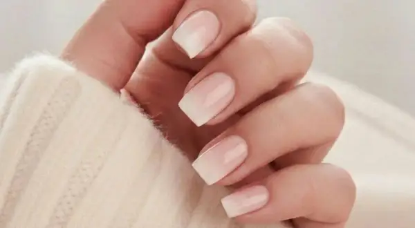 En hand med snygga naglar ombre i en mjuk, ljus ton. Naglarna har en gradvis övergång från en ljusrosa bas till vita toppar, vilket ger en elegant och naturlig look.