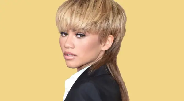 Zendaya visar upp en modern mullet med blont hår och en svart kavaj mot en gul bakgrund.