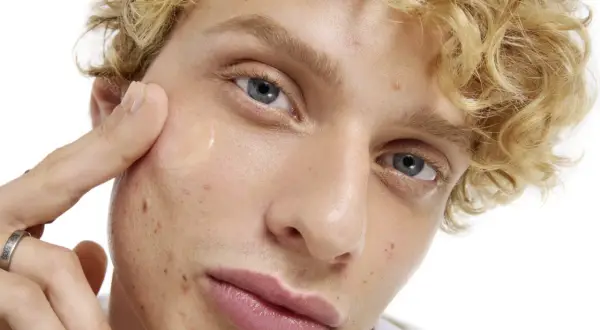 En ung man med lockigt blont hår applicerar en klar gel på kinden, en del av hans resa för att få clear skin.