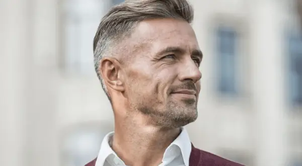 En man med grått hår, klädd i en vit skjorta och en vinröd tröja, tittar bort med ett leende.