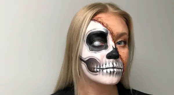 En kvinna med en otrolig ansiktsmålning för halloween. Halva ansiktet är målat som ett skelett med en vit pupill, medan andra halvan visar en realistisk, trasig hudeffekt.