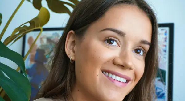 En kvinna ler med en naturlig no makeup makeup-look. En grön Monstera-växt skymtar i förgrunden.