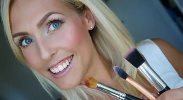 Blond kvinna med blå ögon ler och håller sminkborstar. Hon har precis applicerat rouge för en frisk lyster.