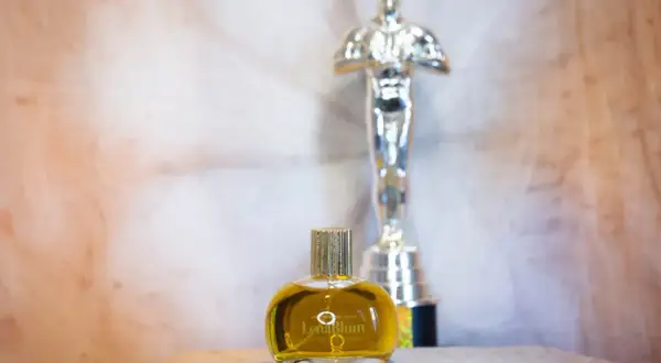 En gyllene flaska med naturliga parfymer, "LenaBlum Out of Woods", står framför en Oscarstatyett.