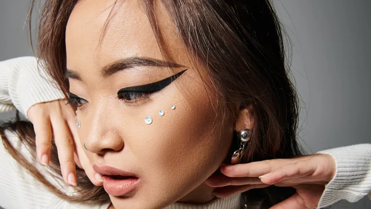 En närbild på en ung kvinna med en dramatisk eyeliner vinge, glittrande kristaller på kinden och en silverfärgad örhänge.