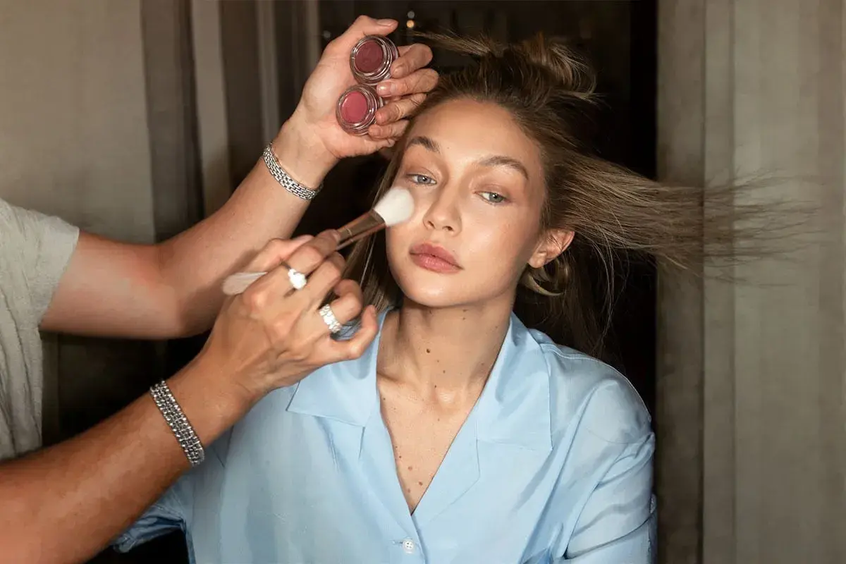 En makeupartist applicerar foundation med en borste på en kvinna med blont hår. Två små burkar med rouge syns i handen.