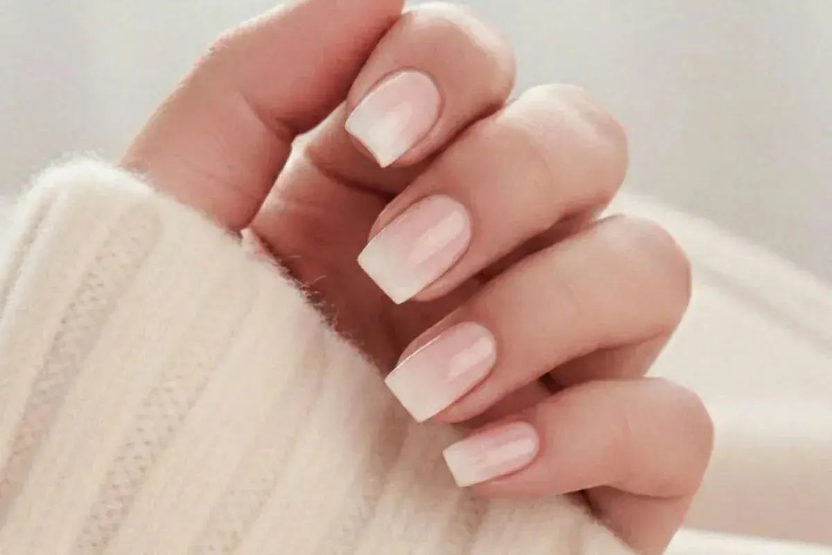 En hand med snygga naglar ombre i en mjuk, ljus ton. Naglarna har en gradvis övergång från en ljusrosa bas till vita toppar, vilket ger en elegant och naturlig look.