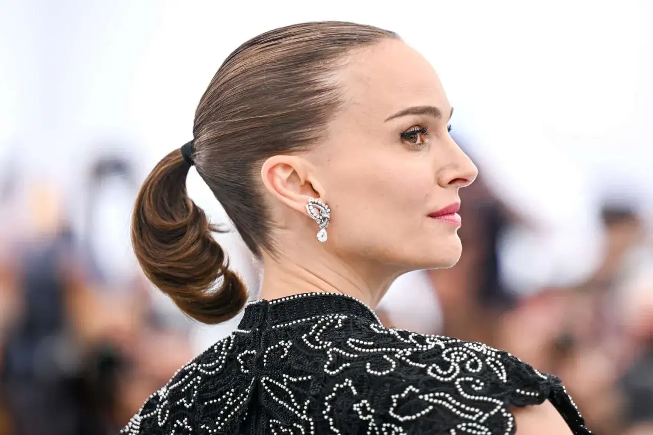 Natalie Portman med en elegant tofs i håret, bär en utsmyckad svart klänning och glittrande örhängen.