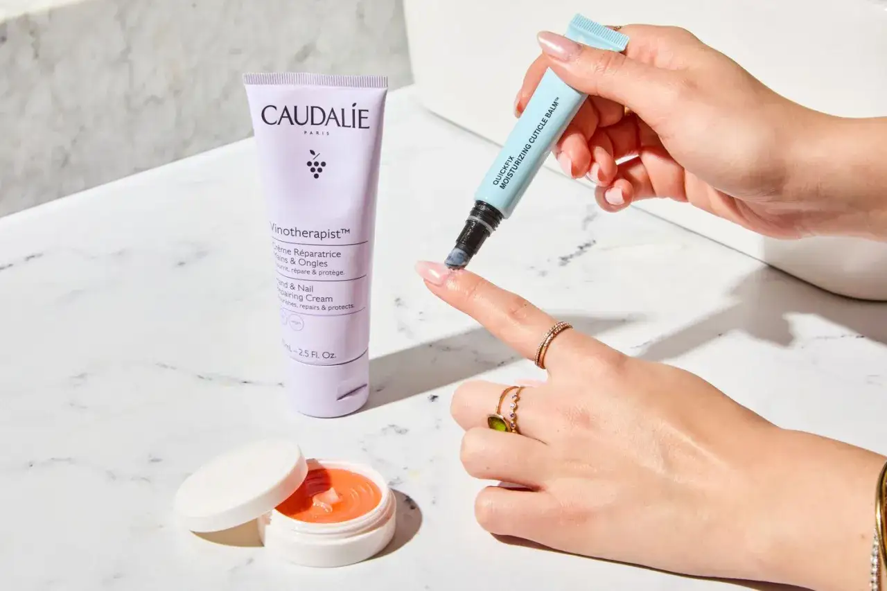 En hand applicerar nagelbandsolja f&ouml;r att motverka torra nagelband. En tub med Caudalie handkr&auml;m syns bredvid.