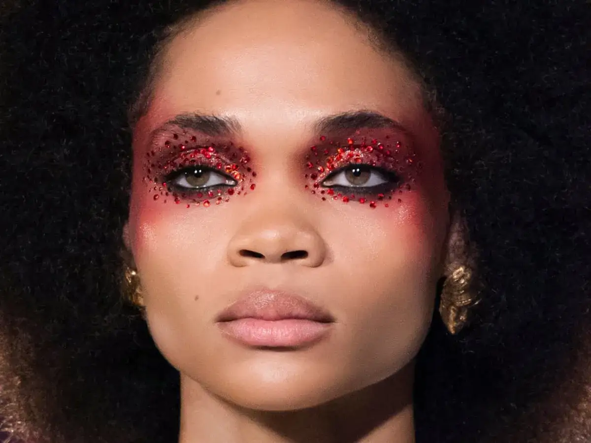 En kvinna med dramatisk &ouml;gonmakeup i r&ouml;tt glitter och guld&ouml;rh&auml;ngen. En doft av amber kan anas i hennes utstr&aring;lning.
