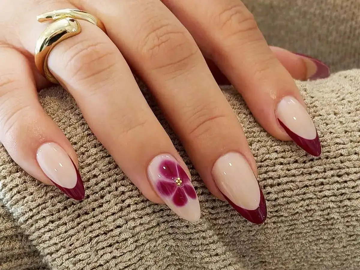 En hand med elegant fransk manikyr i vinrött och beige. En nagel har en vacker blomdekor med gulddetaljer.