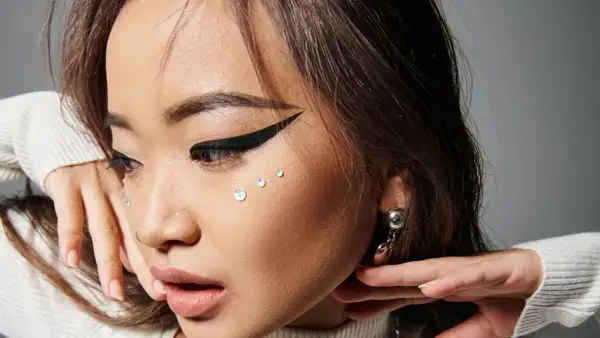 En närbild på en ung kvinna med en dramatisk eyeliner vinge, glittrande kristaller på kinden och en silverfärgad örhänge.