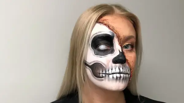 En kvinna med en otrolig ansiktsmålning för halloween. Halva ansiktet är målat som ett skelett med en vit pupill, medan andra halvan visar en realistisk, trasig hudeffekt.