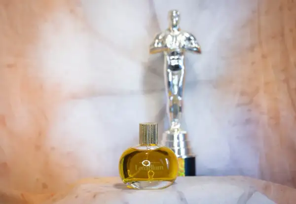 En gyllene flaska med naturliga parfymer, "LenaBlum Out of Woods", står framför en Oscarstatyett.
